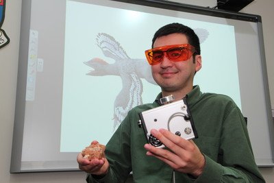 Dr Pittman and the laser-stimulated fluorescence device used for scanning the soft tissues of Archiornis. (Background photo is a life reconstruction of bird-like feathered dinosaur Archiornis)